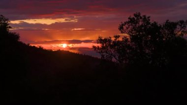 Dramatik bir gün batımı ve ormanlık tepe üzerinde yanan bir gökyüzü. Batan güneş altın ışıklarıyla onların arasından bakarken bulutlar sıcak ve canlı renklerle parıldıyor..