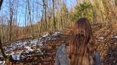 Genç bir kadın köpeğiyle birlikte manzaralı bir sonbahar ormanı patikasında yürüyüş yapıyor, arkadaşlığın ve güzel sonbahar ortamının tadını çıkarıyor. Güneşli bir ormanda ilk kar parçalarıyla yürüyün..