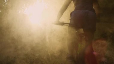 SILHOUETTE, LENS FlaRE: puslu bir sonbahar sabahı erken saatlerde ormanda bisiklet gezisi. Sportif bir kadın bisikletiyle ağaçların arasından süzülen güneş ışınları altında yuvarlanan bir sis bulutunun içinden geçiyor..