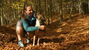 Kadın sonbahar ormanında köpeğinin yanında diz çöker ve köpek onu yalarken gülümser. Sonbahar mevsiminde doğanın rengarenk kucaklamalarında bayanla evcil hayvan arasında nazik bir neşe ve dostluk anı.