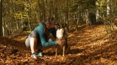 Neşeli kadın, mutlu köpeğini, canlı düşen yapraklarla dolu, güneşli bir ormanda öpüyor. Her ikisi de, sonbahar mevsiminin tadını çıkarırken, altın yapraklar denizinin ortasında sevgi dolu bir anı paylaşıyor..