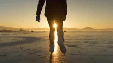 Yavaş hareket, düşük öfke, lensler alev alev, kadın tozluklu buz patenleriyle donmuş gölden altın günbatımına doğru kayıyor. Tanınmayan dişi turist, manzaralı bir kış akşamında buzlu yüzeyde paten kayıyor.
