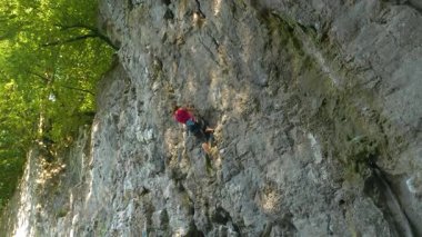 Uçsuz bucaksız bir kadın tırmanışçı kırmızı bir atlet giyerek ve güvenlik teçhizatı kullanarak zorlu bir uçuruma tırmanır. Atletik Kafkasyalı kadın, yazın yemyeşil ormanlarla çevrili dik bir kaya duvarına tırmanıyor..
