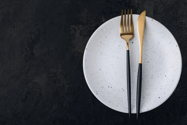Empty plate with fork and knife on dark stone background. Gold and black tableware with white plate, top view, copy space