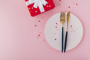 Valentine's Day background. A set of golden cutlery and pink napkin on plate with hearts confetti. Top view, copy space.