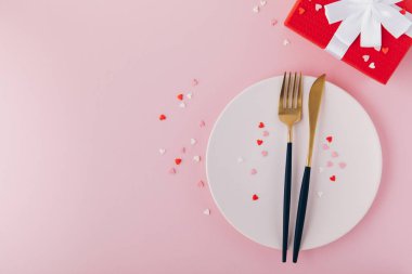 Valentine's Day background. A set of golden cutlery and pink napkin on plate with hearts confetti. Top view, copy space.