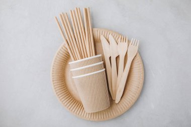 Disposable paper tableware (paper cups, plates, straws, wooden forks, knives, ) on gray stone background, top view, copy space. Plastic free and zero waste concept.