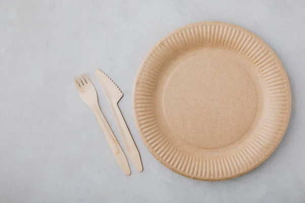 Disposable paper tableware (paper plates, wooden forks, knives, ) on gray stone background, top view, copy space. Plastic free and zero waste concept.
