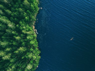 Finlandiya 'da balıkçı teknesi ve yeşil yaz ormanlarıyla mavi su gölünün insansız hava aracı görüntüsü.