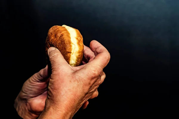An elderly man holds food, a donut, in his hands. Dark background.