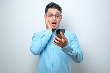 Beyaz stüdyo arka planında iyi haber okumak için akıllı telefon kullanan genç ve yakışıklı bir Asyalı şok oldu.