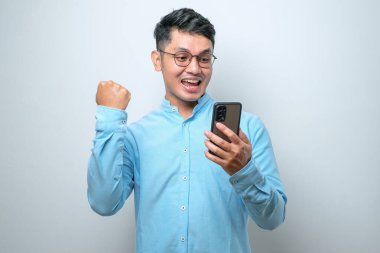 Beyaz stüdyo arka planında iyi haber okumak için akıllı telefon kullanan genç ve yakışıklı bir Asyalı şok oldu.