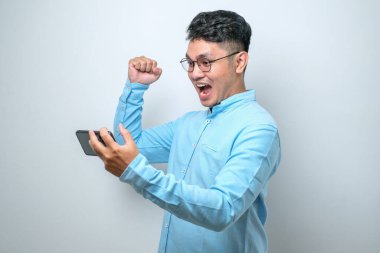 Beyaz stüdyo arka planında iyi haber okumak için akıllı telefon kullanan genç ve yakışıklı bir Asyalı şok oldu.