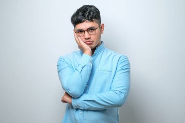 Beyaz arka plan üzerine günlük gömlek ve gözlük takan Asyalı genç adam kollarını kavuşturarak depresyondan sıkılmış ve yorgun görünüyor..