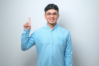 Genç Asyalı bir adam günlük kıyafetler giyer ve beyaz arka planda mutlu ve kendinden emin gülümserken bir numaralı parmaklarıyla işaret eder.