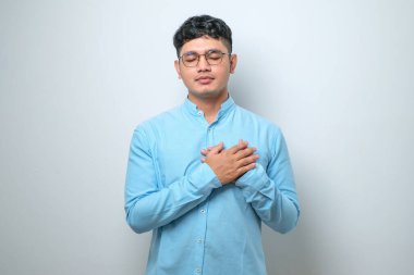 Yakışıklı, gündelik kıyafetler giyen, elleri göğsünde, gözleri kapalı gülümseyen ve beyaz arka planda minnet dolu bir jest yapan genç adam. sağlık kavramı.