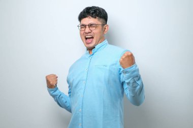 Asyalı genç bir adam, izole edilmiş beyaz arka planda dikilmiş mutlu ve heyecanlı bir şekilde kollarını kaldırıp, gülümseyerek ve başarı çığlıkları atarak zafer jesti yapıyor. Kutlama konsepti