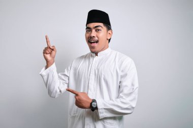 Beyaz pıhtılaşmış ve kafatası şapkalı gülümseyen Asyalı genç Müslüman adam beyaz arka planda izole edilmiş elini kullanarak boşluğu kopyalamak için üst solu işaret ediyor.