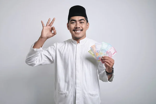 Smiling asian muslim man in white clothes and black skull cap smiling happy while holding paper money and showing okay gesture using hand isolated over white background