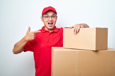 Kırmızı üniformalı kendine güvenen Asyalı kurye çağrı servisini teşvik ediyor, beyaz arka planda izole edilmiş bir şekilde karton kutuyu gösteriyor ve işaret ediyor.