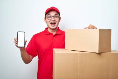 Kırmızı üniformalı Asyalı kurye, beyaz arka plan üzerinden izole edilmiş, boş ekranlı akıllı telefonu stüdyoda tutuyor.