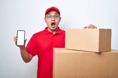 Kırmızı üniformalı Asyalı kurye, beyaz arka plan üzerinden izole edilmiş, boş ekranlı akıllı telefonu stüdyoda tutuyor.