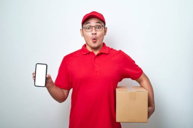 Kırmızı üniformalı boş telefon ekranı gösteren mutlu Asyalı teslimatçı beyaz arka planda izole edilmiş karton kutu tutuyor..