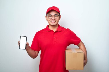Kırmızı üniformalı boş telefon ekranı gösteren mutlu Asyalı teslimatçı beyaz arka planda izole edilmiş karton kutu tutuyor..