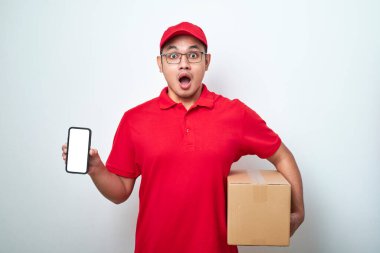 Kırmızı üniformalı boş telefon ekranı gösteren mutlu Asyalı teslimatçı beyaz arka planda izole edilmiş karton kutu tutuyor..