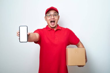 Kırmızı üniformalı boş telefon ekranı gösteren mutlu Asyalı teslimatçı beyaz arka planda izole edilmiş karton kutu tutuyor..