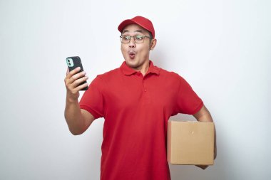 Kırmızı üniformalı Asyalı kurye, beyaz arka planda izole edilmiş karton kutuyu tutarken akıllı telefonuna bak.