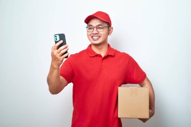 Kırmızı üniformalı Asyalı kurye, beyaz arka planda izole edilmiş karton kutuyu tutarken akıllı telefonuna bak.