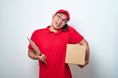 Gülümseyen Asyalı kurye telefona cevap veriyor, cep telefonunu omzunda tutuyor, müşteriye teslimat için paketi hazırlıyor ve not alıyor, müşteriyle telefon görüşmesi yapıyor, beyaz bir geçmişi var.