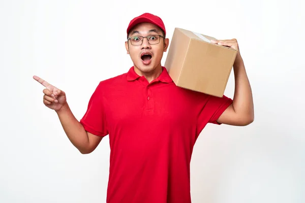 stock image Suprised Asian courier in red shirt and cap holding box package on shoulder and point left, isolated over white background