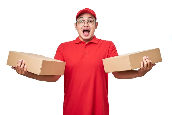 stock image Surprised asian courier man wearing red uniform standing over white background holding two boxes with orders, packing shipping items for clients.