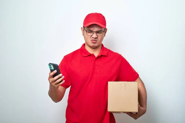 Angry asian courier man in red uniform look at his smartphone while holding cardboard box parcel isolated over white background