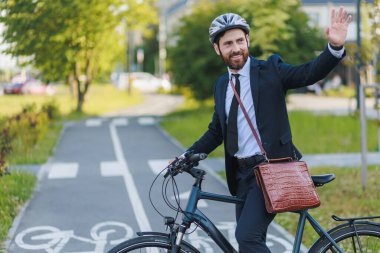 Smiling male manager saying goodbye after work, while getting to bike on street. Side view of happy boss in formal apparel, standing on cycle path and waving hand to coworkers. Concept of gesturing.