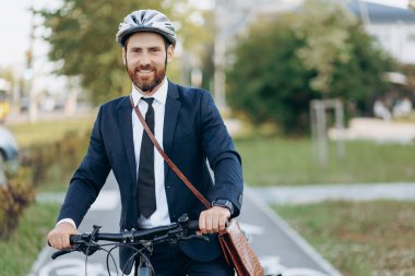 Elegant businessman in protective helmet looking at camera, smiling, while cycling to work outdoors. Portrait of happy executive using eco transport, while commuting to job. Concept of eco lifestyle.