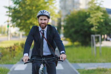 Smiling male employee in black suit commuting to work on bike lane in sunny morning. Front view of happy bearded worker looking at camera, while driving bicycle outdoors. Concept of eco lifestyle.