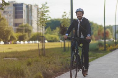 Joyful man in black suit riding on bike on paved road after workday in summer. Front view of smiling businessman wearing smart casual cycling through sleeping area of town. Concept of eco commute.