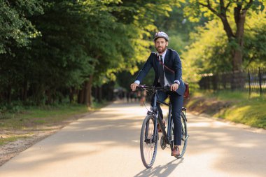 Smiling bearded executive in business apparel returning from job on bike through green area. Front view of joyful office clerk feeling great, while cycling home in sunny day. Concept of eco lifestyle.