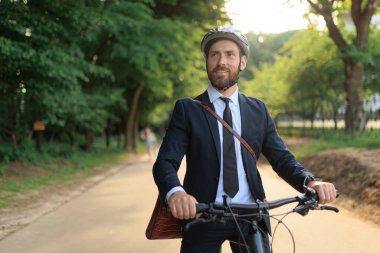 Successful entrepreneur enjoying view, while riding on bike to work on park alley. Portrait of attractive bearded man handle steering, while cycling to business meeting at day. Concept of eco vehicle.