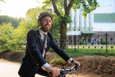 Happy male manager in black suit, riding on bicycle after workday in city. Side view of smiling worker in helmet looking ahead, enjoying trip, while driving bike outdoors. Eco friendly concept.