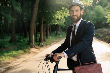 Confident businessman in helmet walking to work with bicycle in morning. Portrait of bearded entrepreneur in stylish suit standing with bike, while looking at camera in town. Concept of lifestyle.