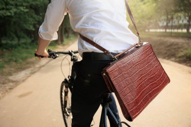 Crop view of anonymous office clerk, wearing white shirt and brown leather suitcase, getting to work on bike against sunlight. Back view of male stylish businessman cycling in morning. Work concept.