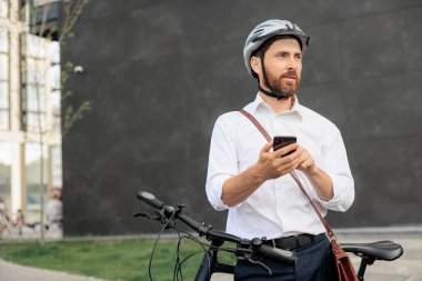 Bearded man surfing online map in smartphone, while searching way to job in morning. Front view of concentrated male employee in white shirt relying on parked bike, while making call. Concept of work.