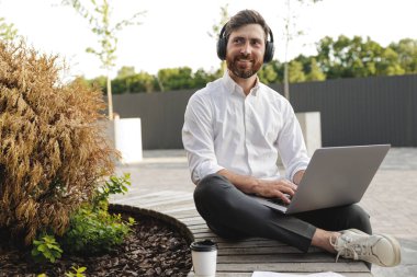 Joyful freelancer sitting on wooden bench with portable laptop on knees. Handsome man in black headphones looking aside with smile on face while working outside.