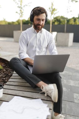 Positive office worker in headphones looking on laptop screen with joyful facial expression. Stylish male working on computer while sitting on wooden bench with leg.