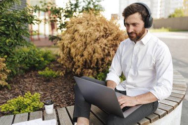 Handsome bearded businessman in formal clothing typing on portable laptop. Positive male in headphones attentively looking on screen while sitting on bench in urban area.