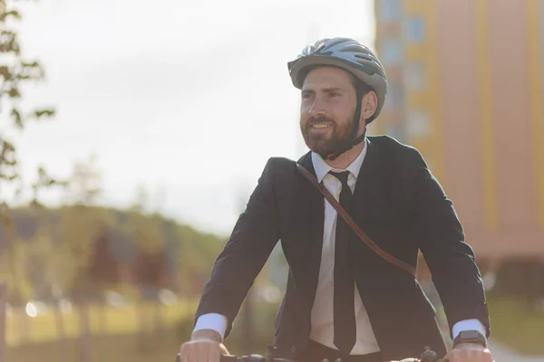 Happy bearded male businessman in protective helmet commuting to job place. Portrait of young cheerful man in suit and tie looking around, while riding, with blurred background. Concept of lifestyle.